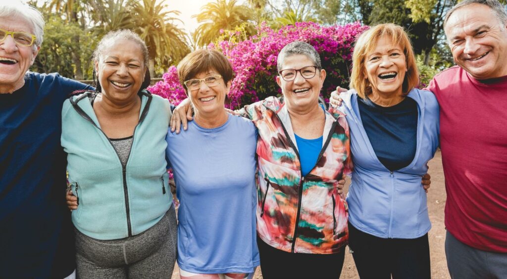 A group of seniors with arms around each others' shoulders smiling in an outdoor garden.