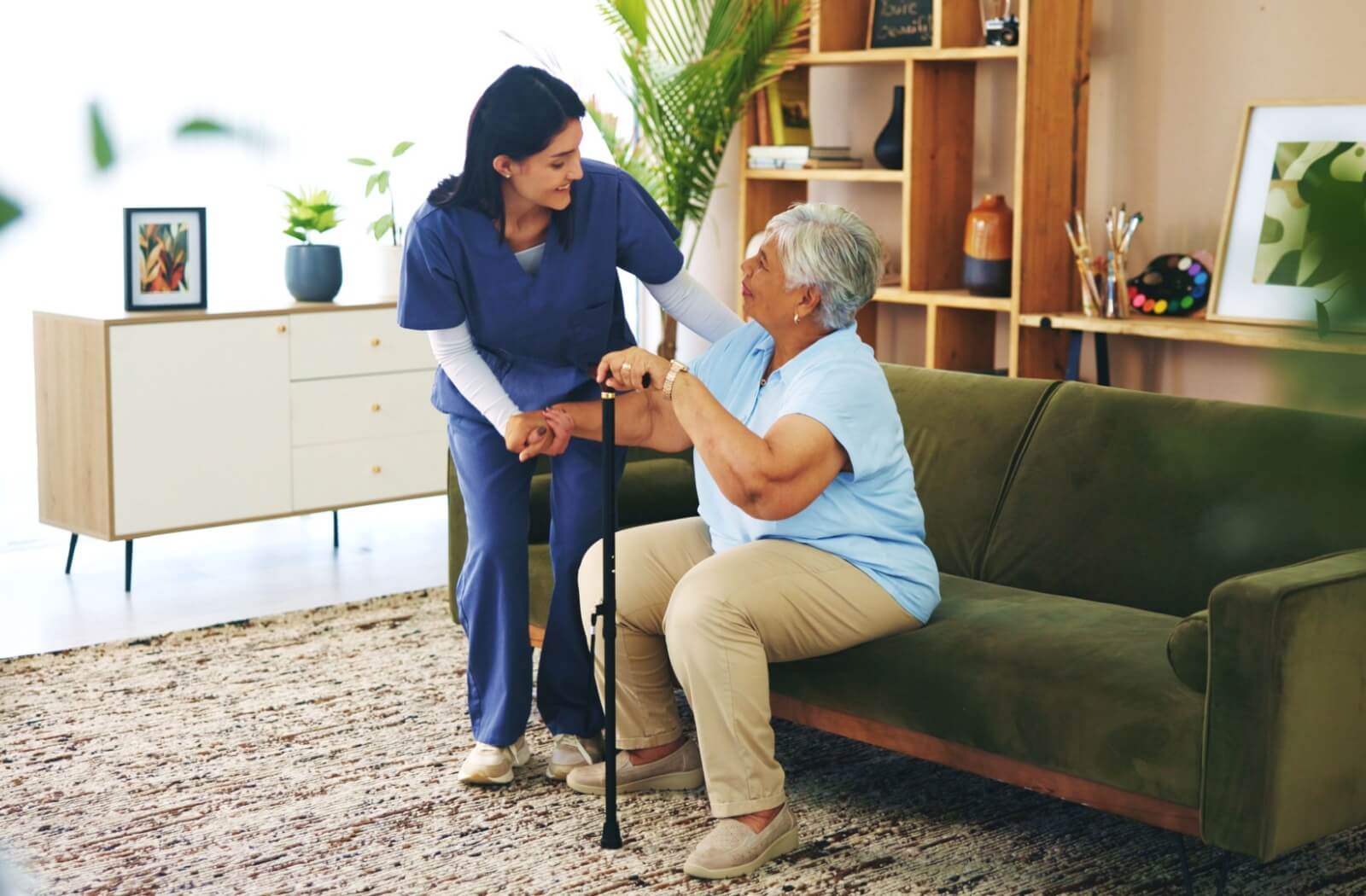 Caregiver assisting a seated person with a cane in a bright living room decorated with artwork, plants, and wooden shelves