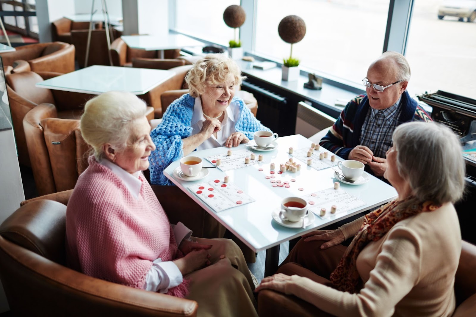 Four seniors sit at a table, smiling as they play bingo and enjoy tea together.